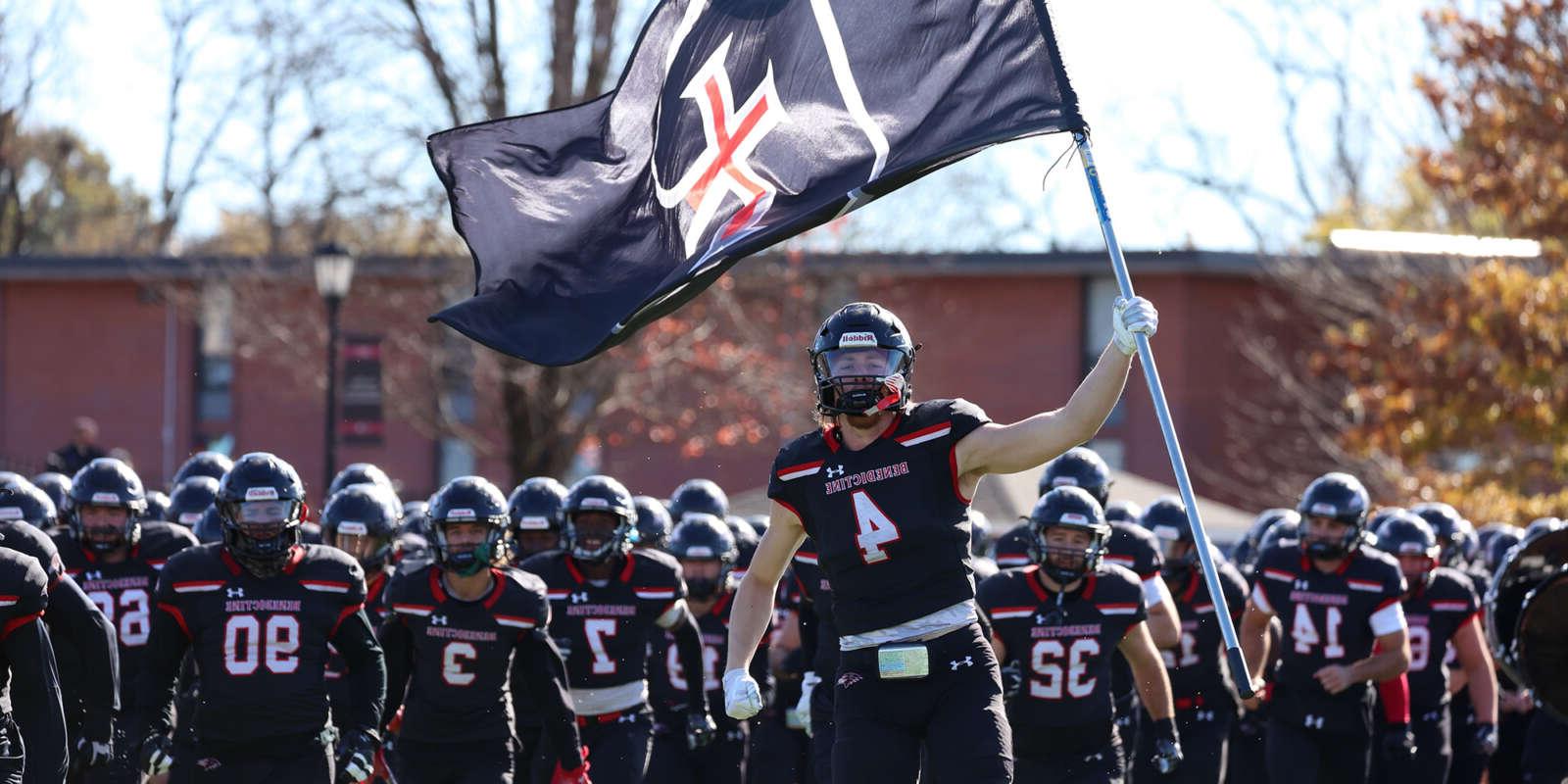 The Raven Football team runs onto the field, with one player holding the Benedictine College flag